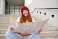 Young smiling redhead girl, tourist sits on stairs outdoors with city paper map, looking for direction, traveller Royalty Free Stock Photo