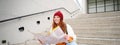 Young smiling redhead girl, tourist sits on stairs outdoors with city paper map, looking for direction, traveller Royalty Free Stock Photo