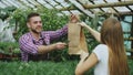 Young smiling seller florist working in garden center. Man gives shopping bag to customer and making payment by credit