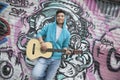 Young smiling street musician leaning on a wall with graffiti drawings and playing his guitar