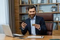 Young smiling and successful male businessman sitting in office at desk and using mobile phone and credit card Royalty Free Stock Photo