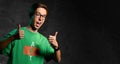 Young smiling teen boy in green t-shirt, jeans and glasses standing and showing good thumbs sign over dark background Royalty Free Stock Photo