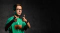 Young smiling teen boy in green t-shirt, jeans and glasses standing and showing good thumbs sign over dark background Royalty Free Stock Photo