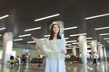 Young smiling traveler tourist woman in hat holding paper map, looking aside while waiting in lobby hall at Royalty Free Stock Photo