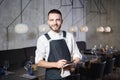 A young, smiling waiter in a restaurant, standing next to the tables with a glass of wine. Dressed in an apron, will take an order