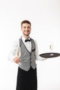 Young smiling waiter in uniform joyfully holding tray while offering glass of champagne on camera over white background Royalty Free Stock Photo