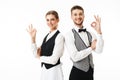 Young smiling waiter and waitress in white shirts and vests sstanding back to back happily looking in camera while