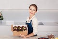 Young smiling woman baker in apron holding paper delivery packaging box with just cooked cupcakes or muffins