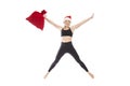 Young smiling woman in a black tracksuit in a Santa Claus hat and a red bag with gifts is jumping. Full height. Isolated over