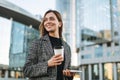 Young smiling woman in coat with coffee cup using mobile phone in evening city street