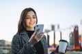 Young smiling woman in coat with coffee cup using mobile phone in evening city street