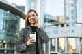 Young smiling woman in coat with coffee cup using mobile phone in evening city street