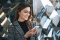 Young smiling woman in coat with coffee cup using mobile phone in evening city street