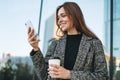 Young smiling woman in coat with coffee cup using mobile phone in evening city street