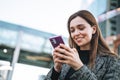 Young smiling woman in coat using mobile phone in evening city street