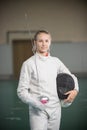 A young smiling woman fencer standing in the gym holding sword and helmet