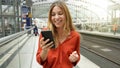 Young smiling woman holding mobile phone on sun flare platform station. Traveler girl on train station using cellphone. Digital Royalty Free Stock Photo