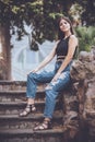 Young smiling woman jeans and accessories. Outdoors sitting on a low wall of rock on a staircase. Long hair
