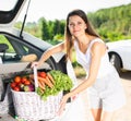 Young smiling woman puts basket of vegetables in the trunk of car