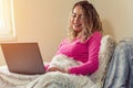 Young smiling woman is relaxing in bed and surfing the net on laptop