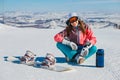 A young smiling woman sits on a mountain slope with a snowboard and a thermos Royalty Free Stock Photo