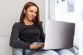 Young smiling woman sitting on modern chair near the window in light cozy room at home working on laptop in relaxing atmosphere Royalty Free Stock Photo