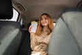Young smiling woman sitting in the rear passenger seat and holding a cup of hot drink while waiting in a traffic jam
