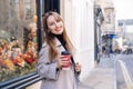 Young smiling woman in trench coat enjoying hot coffee drink in reusable cup while walking on the city streets decorated