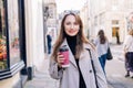 Young smiling woman in trench coat enjoying hot coffee drink in reusable cup while walking on the city streets decorated