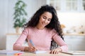 Young Smiling Woman Writing Notes In Notepad, Sitting At Table In Kitchen Royalty Free Stock Photo