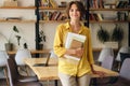 Young smiling woman in yellow shirt leaning on desk with notepad and papers in hand while happily looking in camera in Royalty Free Stock Photo