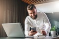 Young smilling bearded businessman standing near table in front of laptop, using smartphone. Man checks e-mail Royalty Free Stock Photo