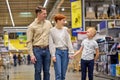 Young Son Child Boy Walking With Parents In Supermarket While They Holding Hands Looking For New Things For Home