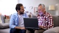 Young son explaining aged father how to use laptop, modern technologies, device