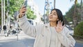 A young south asian woman takes a selfie on a sunny city street, waving at her phone with a smile