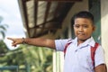 A young southeast asian school boy points to the left while outside a small rural school building