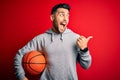 Young sports man holding basketball ball over red isolated background pointing and showing with thumb up to the side with happy Royalty Free Stock Photo