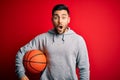 Young sports man holding basketball ball over red isolated background scared in shock with a surprise face, afraid and excited