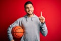 Young sports man holding basketball ball over red isolated background surprised with an idea or question pointing finger with