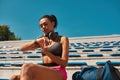 Young sportswoman checking results after training using smartwatch while resting on the stadium seat outdoors Royalty Free Stock Photo