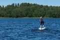 A young sporty blonde woman in the wetsuit is surfing on the sup board in the aquatory of the forest lake on bright summer day.