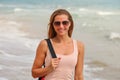 Young sporty brunette woman in sunglasses, standing on the beach, blued sea behind she, sun slightly shining at her. Detail to