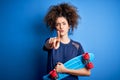 Young sporty woman with curly hair and piercing holding skate over blue background pointing with finger to the camera and to you, Royalty Free Stock Photo