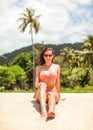 Young sporty woman in orange bikini and sunglasses sits on fine beach sand, wind in her hair, palm tree and jungle behind her Royalty Free Stock Photo