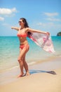 Young sporty woman in red bikini and sunglasses, stand on the beach, holds pink scarf waving in wind behind her. Turquoise sea and