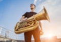 Young street musician playing tuba at the sunset sky background.