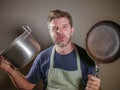 Young stressed and funny lazy man with apron holding kitchen pan and kitchen pot in stress desperate crying isolated background