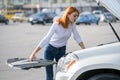 Young stressed woman driver standing near broked car with open hood