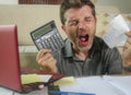 Young stressed and worried man at home living room using calculator and laptop doing domestic accounting paperwork screaming crazy