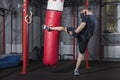 Young strong mixed martial arts fighter delivers strong side kick to punching bag. Active exercise in gym.